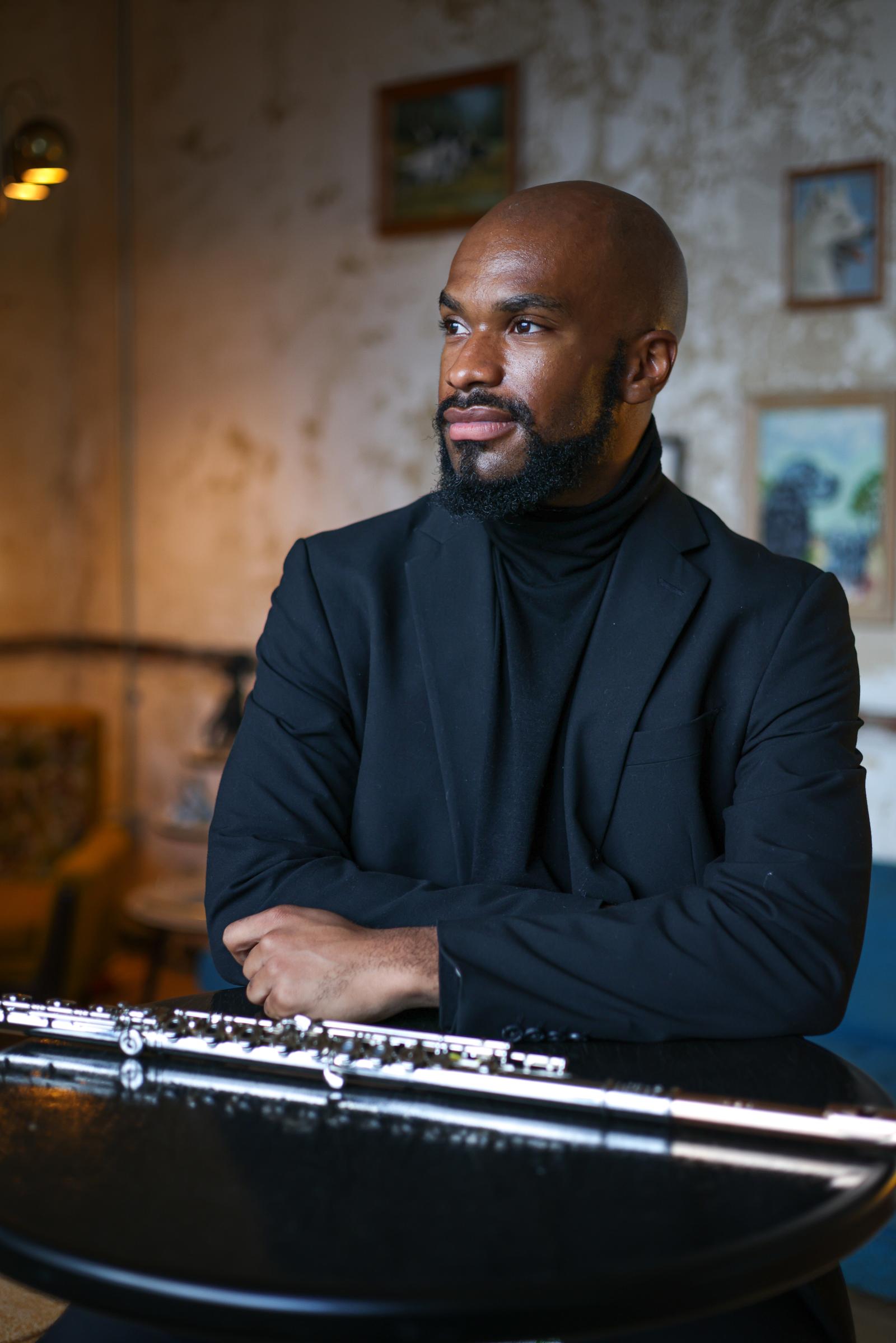 Musician Adam Sadberry sits at a table wearing a black suit, looking to the left of the photo with his flute on the table in front of him.