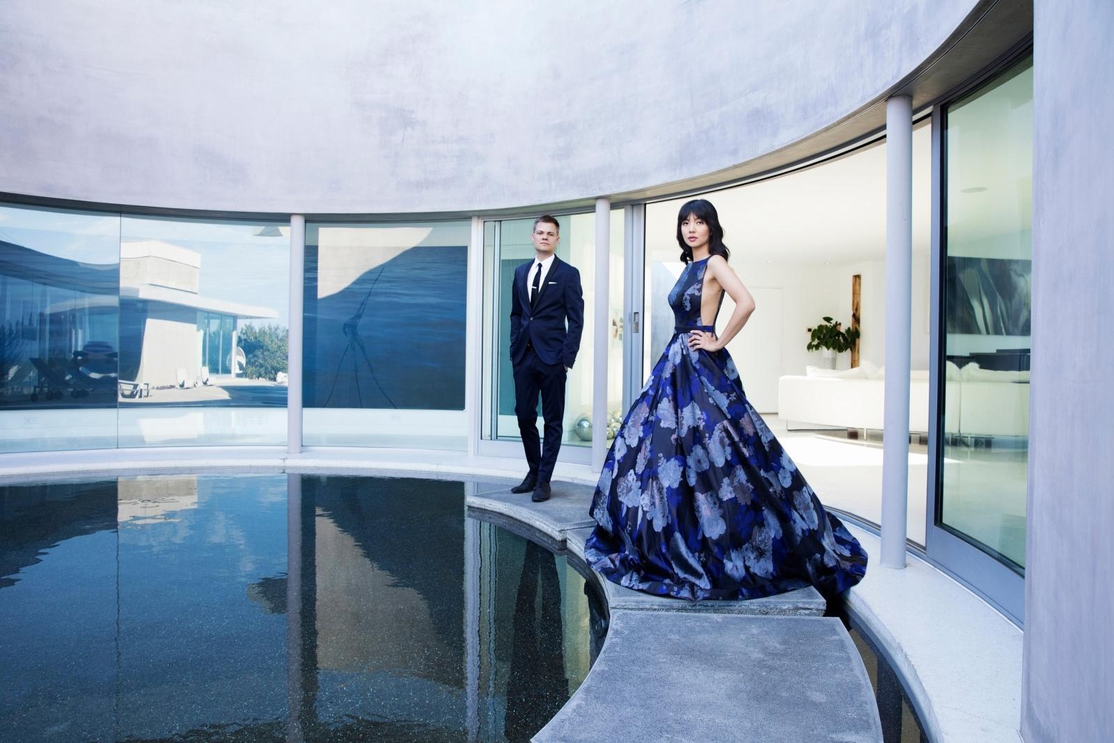 Anderson and Roe stand at the edge of a still reflecting pool a row of curved windows behind them. 
