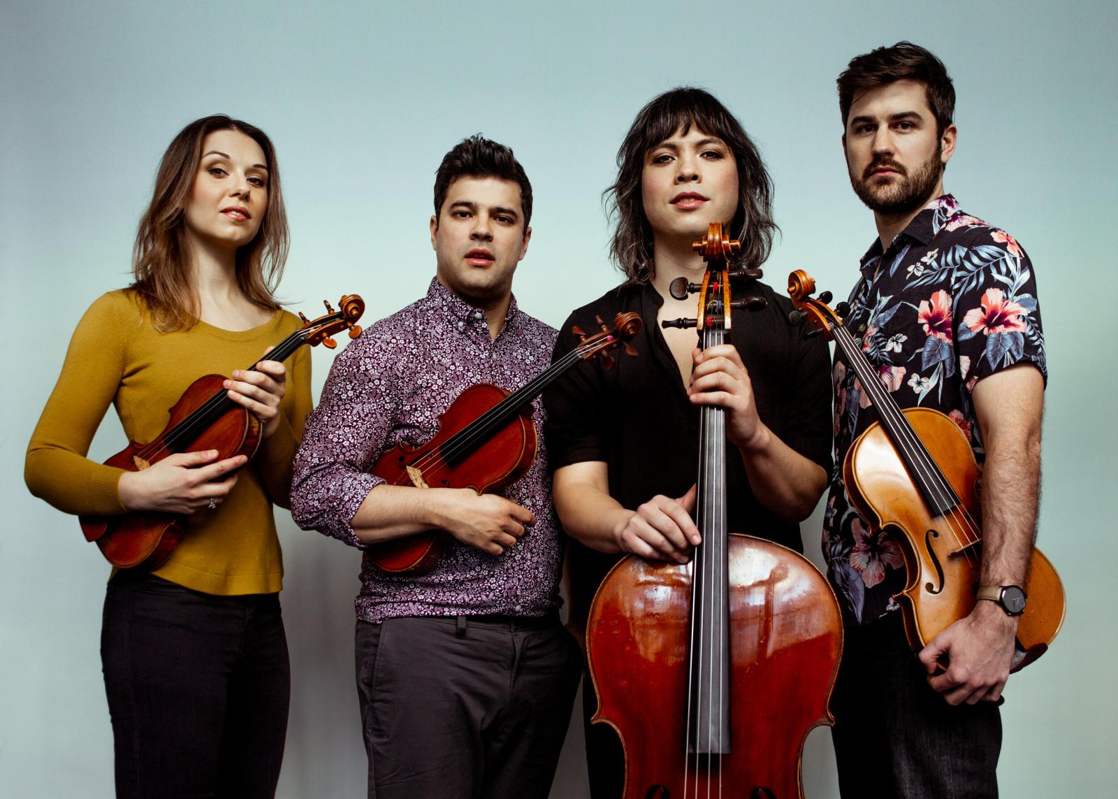 Members of the Attacca string quartet stand with their instruments in front of a light blue background.