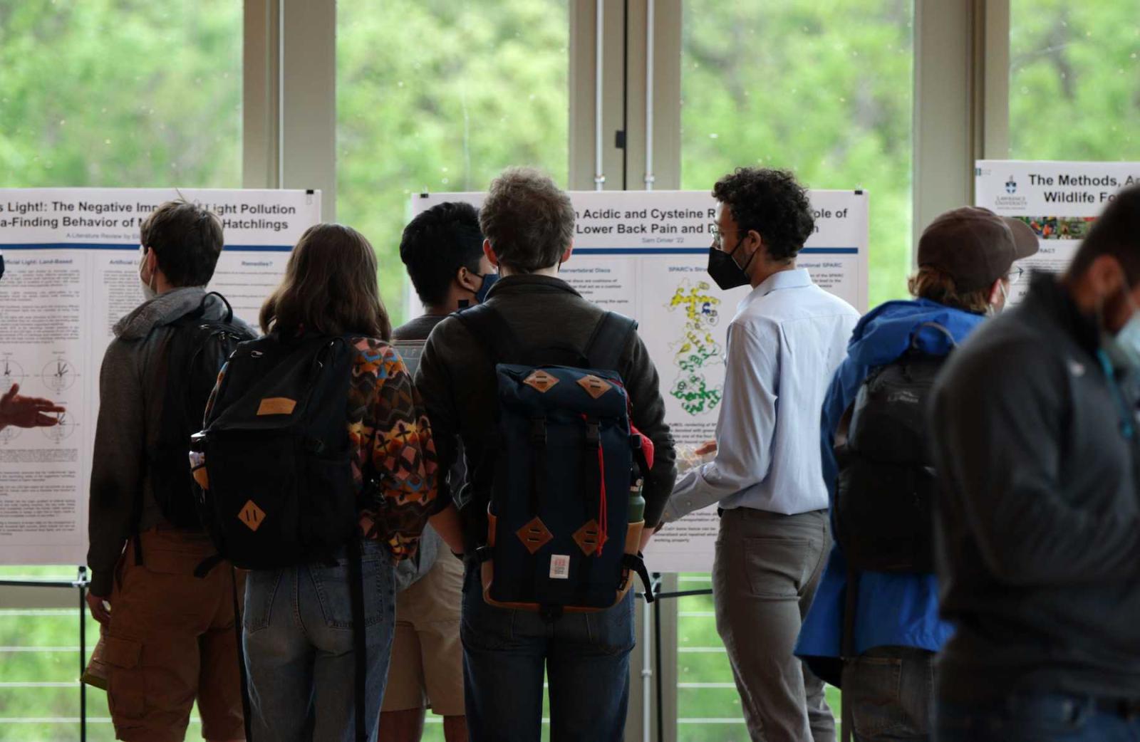 Visitors to Biofest gather around students presenting their work on standing white boards.