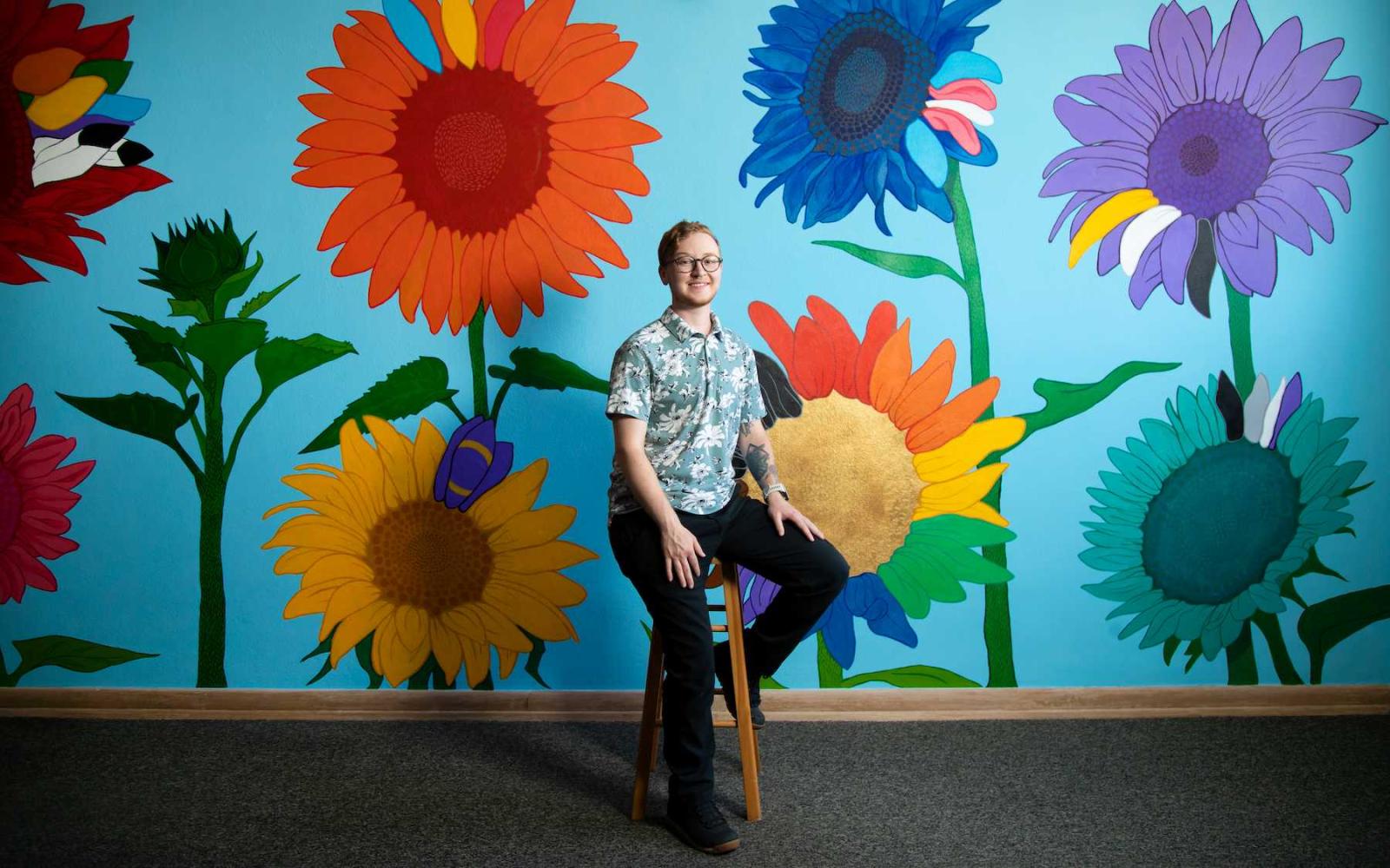 Cael Neary sits on a stool in front of his colorful mural in the Gender and Sexuality Diversity Center.