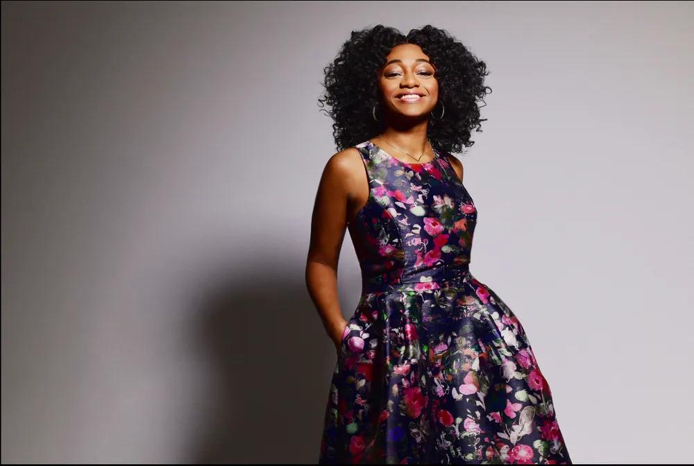 Singer Samara joy smiles at the camera wearing a floral ball gown in front of a grey background. 