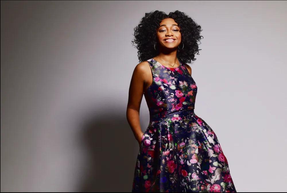 Singer Samara joy smiles at the camera wearing a floral ball gown in front of a grey background.