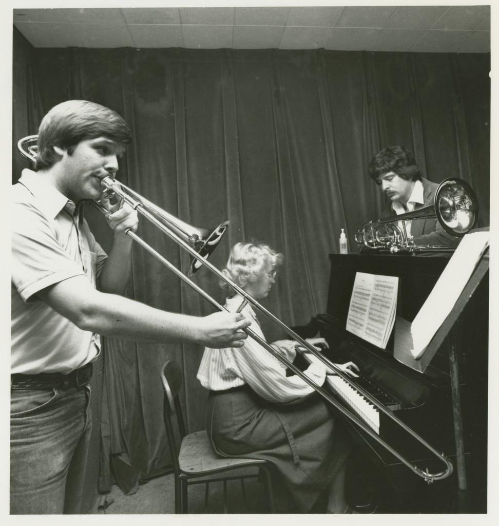A trombone player and a pianist play while a man leans on the paino.