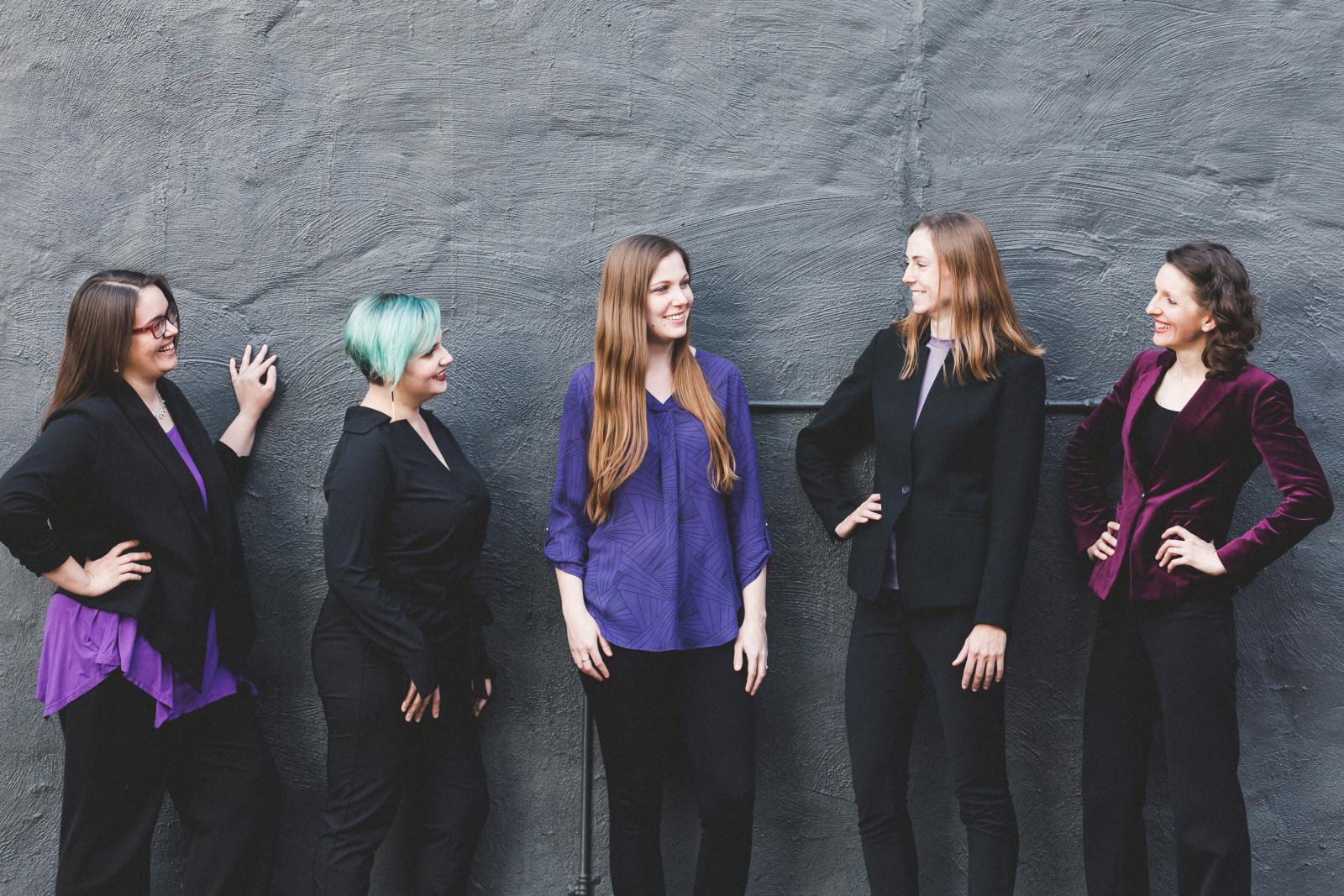 Five women standing against a wall, smiling and looking at each other.