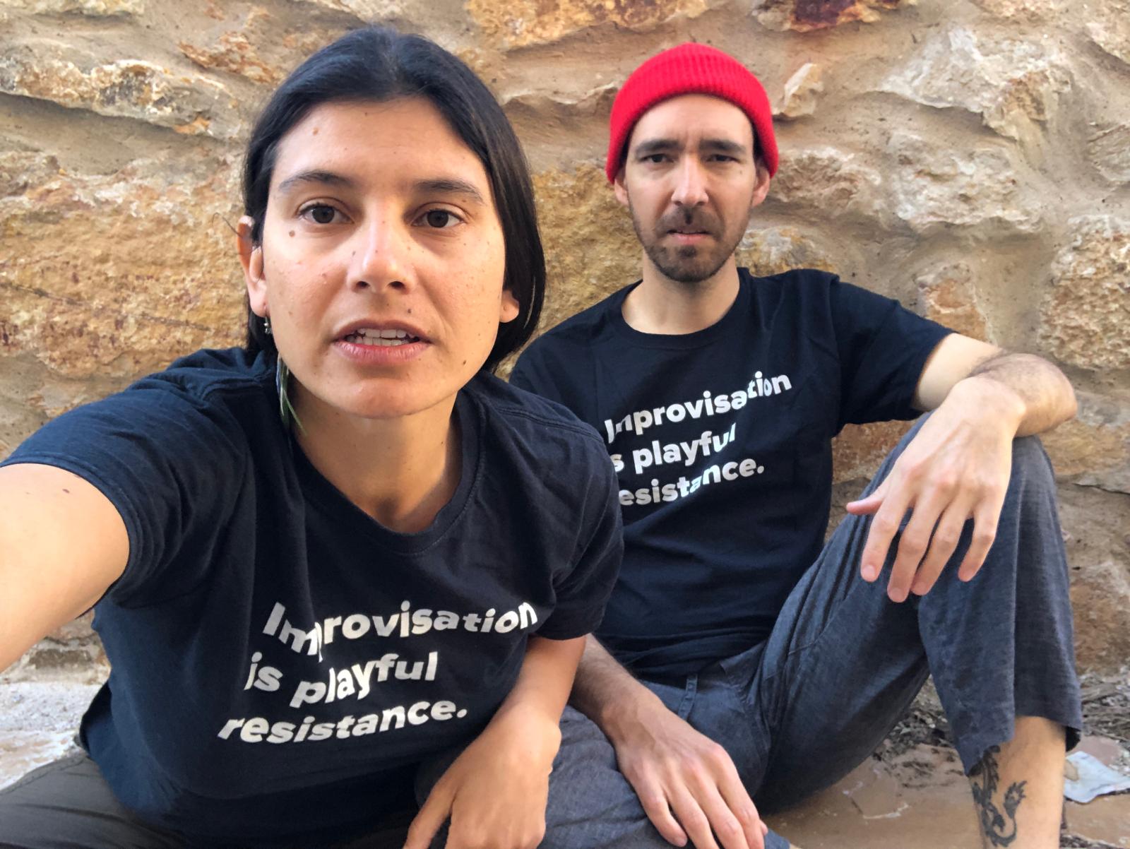 A woman and a man seated against a rocky wall, both wearing T-shirts with the text "Improvisation is playful resistance".