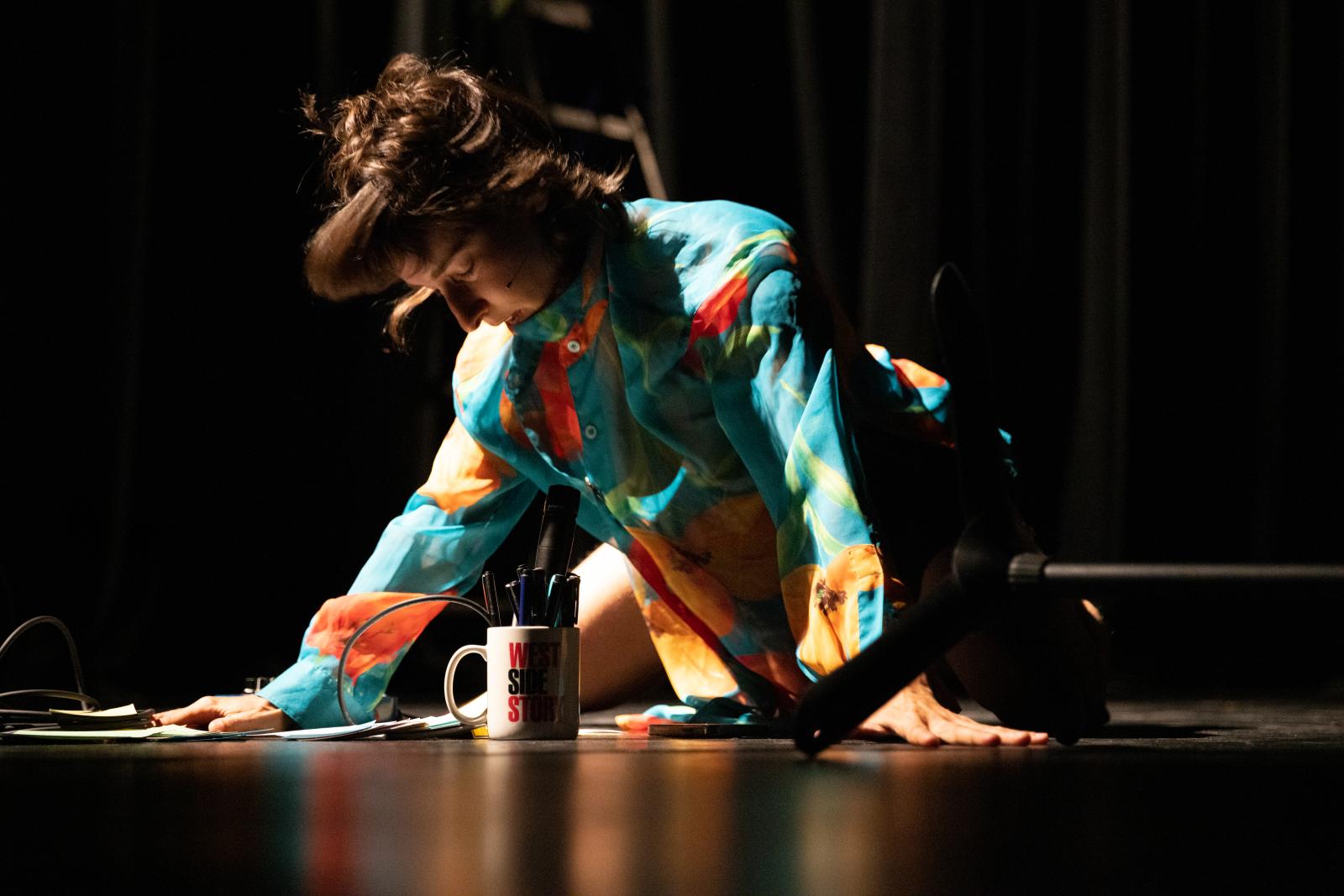 A woman kneeling on the floor, with papers and a cup full of pens before her.