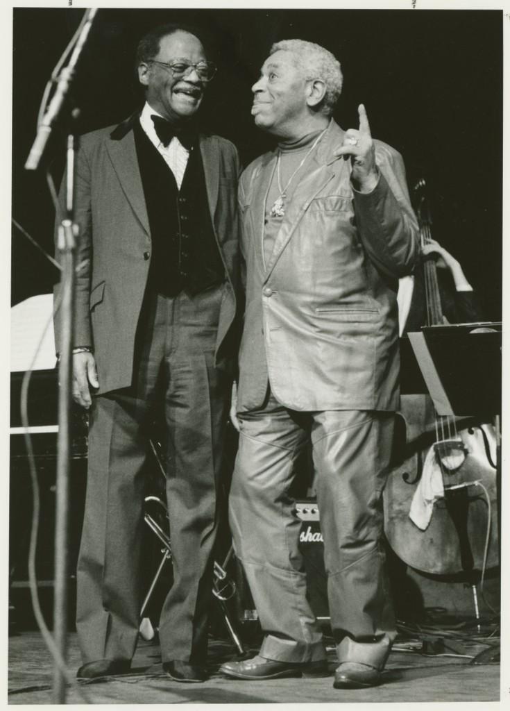 Two men in concert attire stand on a stage.