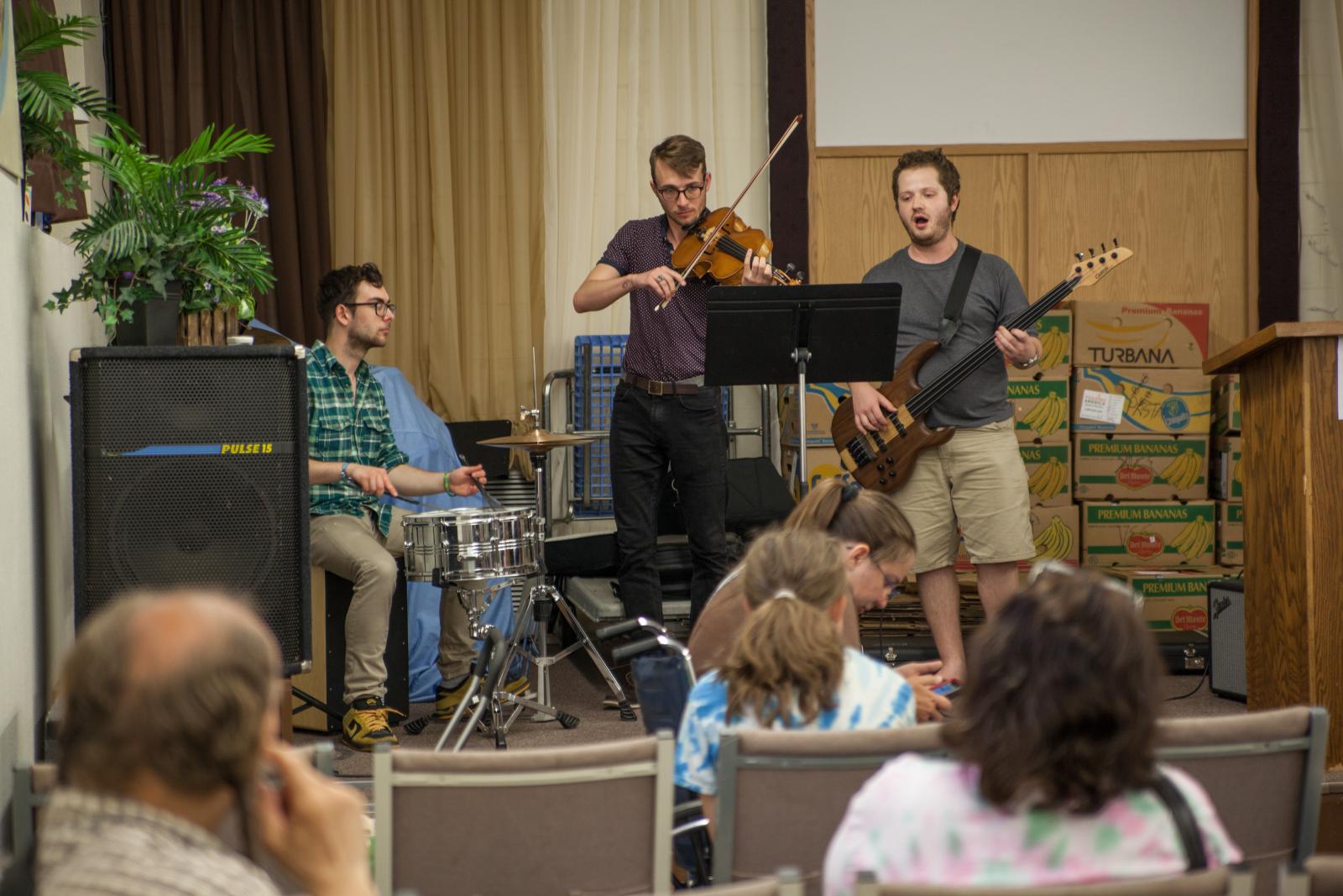 Drummer, violinist, and guitarist perorm on a small stage.