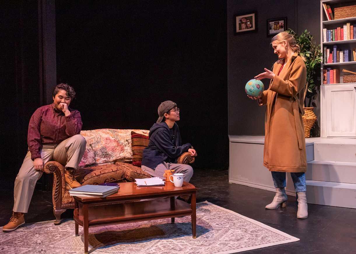 2 seated actors and 1 standing, having a conversation in a living room