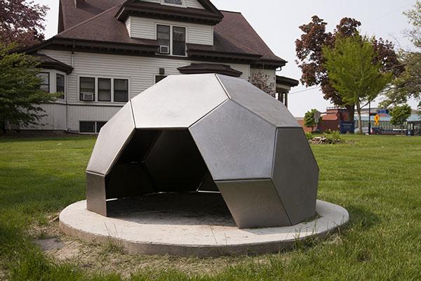 A silver dome made of 5 sided panels with an opening in the front, on a circular concrete pad surrounded by lawn.