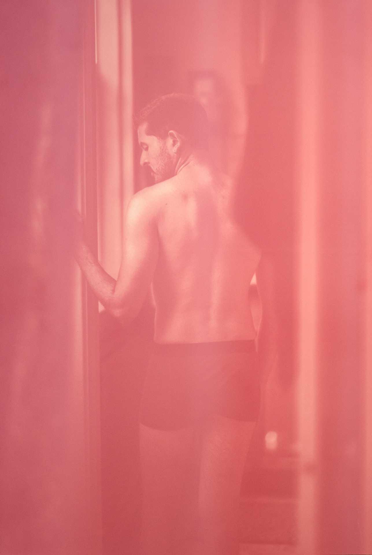 A man in underwear, seen from behind, glances back and leans one arm against a wall; the image has a pink tint.