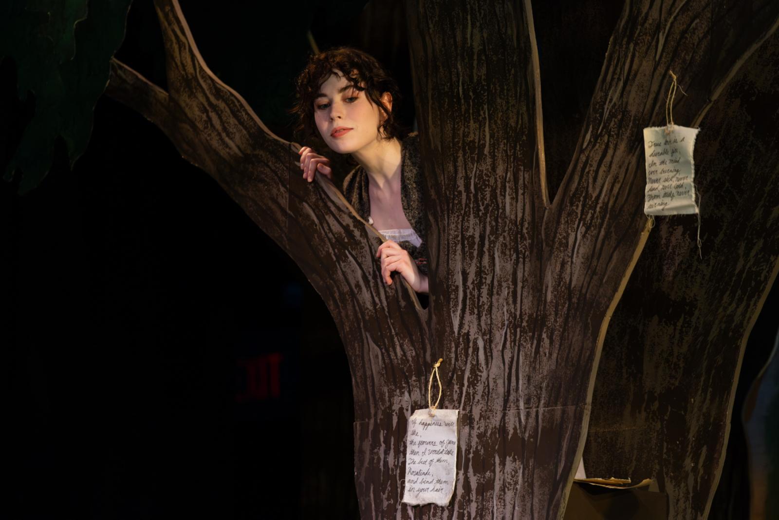 A cast member looks on from a tree on stage.