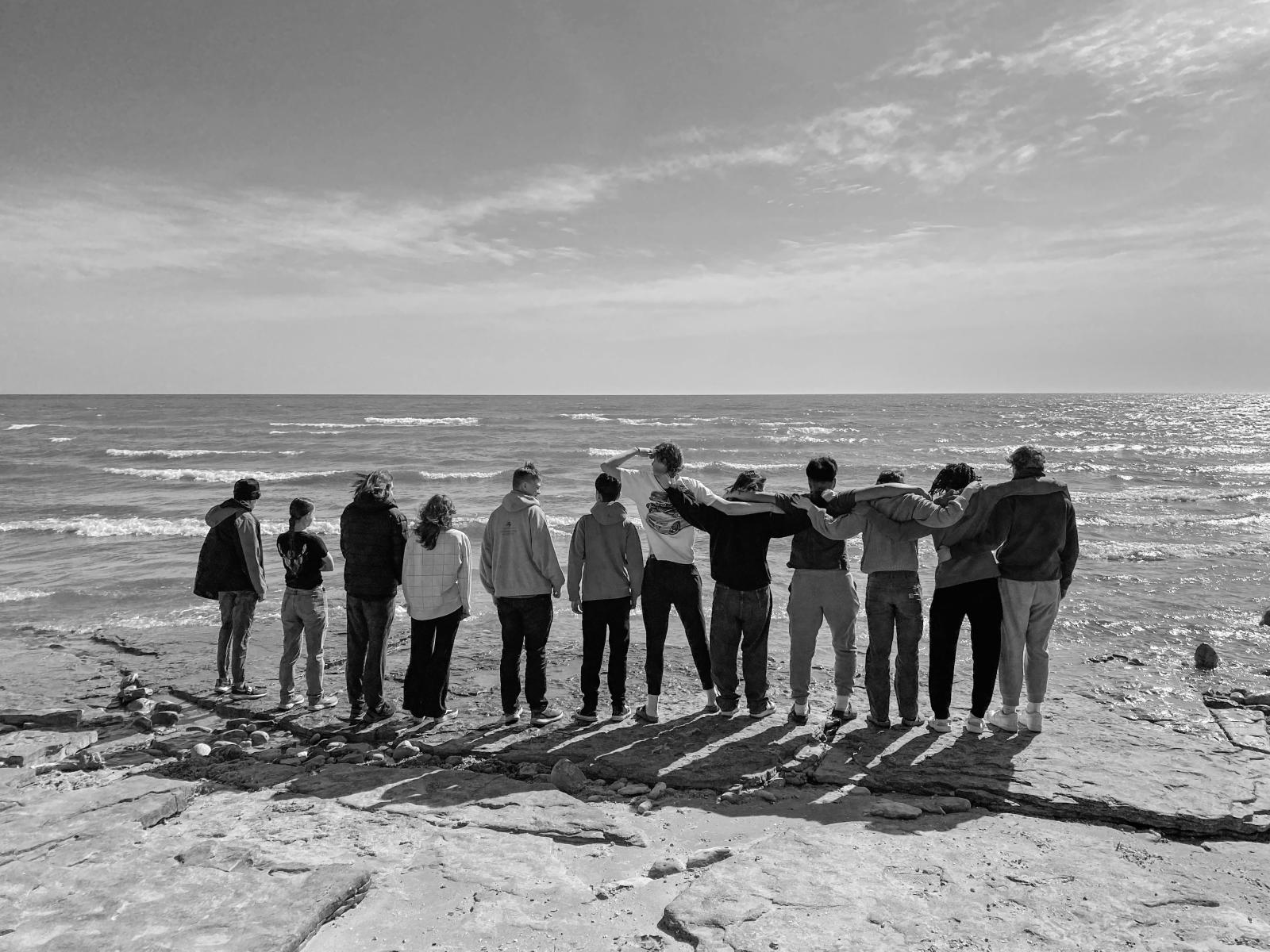 A group of students stand with their backs facing the camera on a rocky shoreline