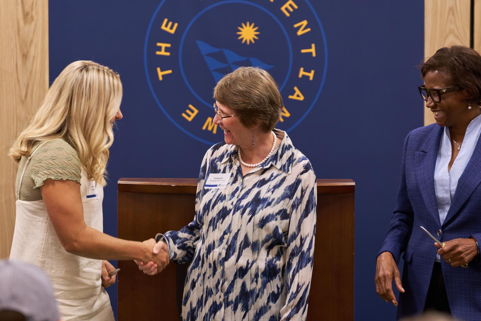 Ratnibd H. Herzog Proffesor of Science and professor of biology, Beth De Stasio shaking hands with neuroscience major, Class of 2026, Taylor Svoboda. President Laurie Carter smiling...