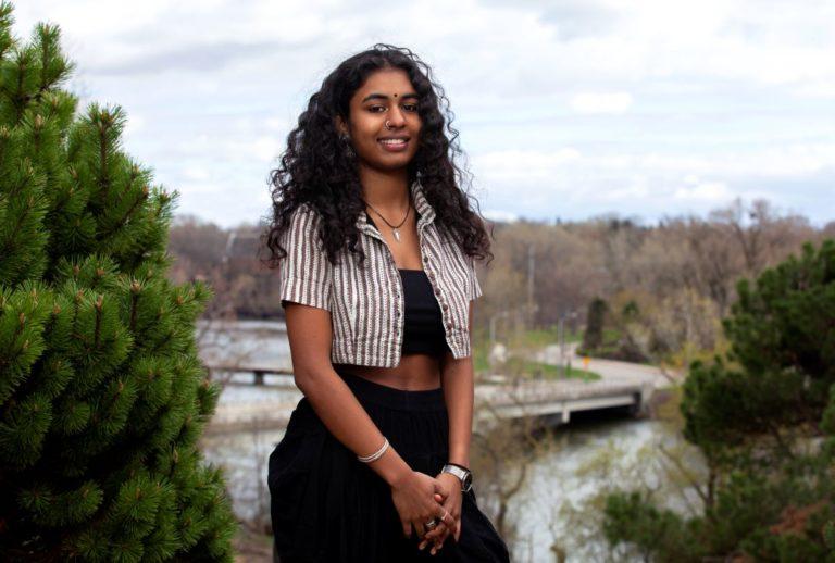 Adya Kadambari, wearing a white and grey striped shirt, smiles at the camera.