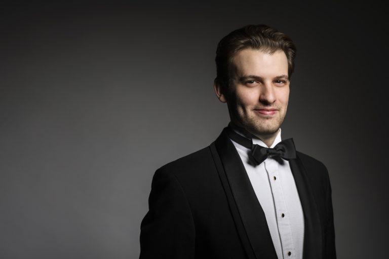 Evan Bravos, wearing a tuxedo, smiles at the camera against a black background.