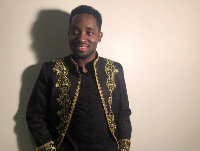 Louric Rankine, wearing a black and gold jacket, stands in front of a white wall and smiles.