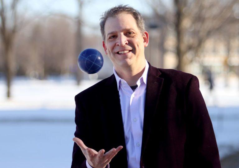 David Gerard, wearing a black suit jacket, throws a blue ball in the air.