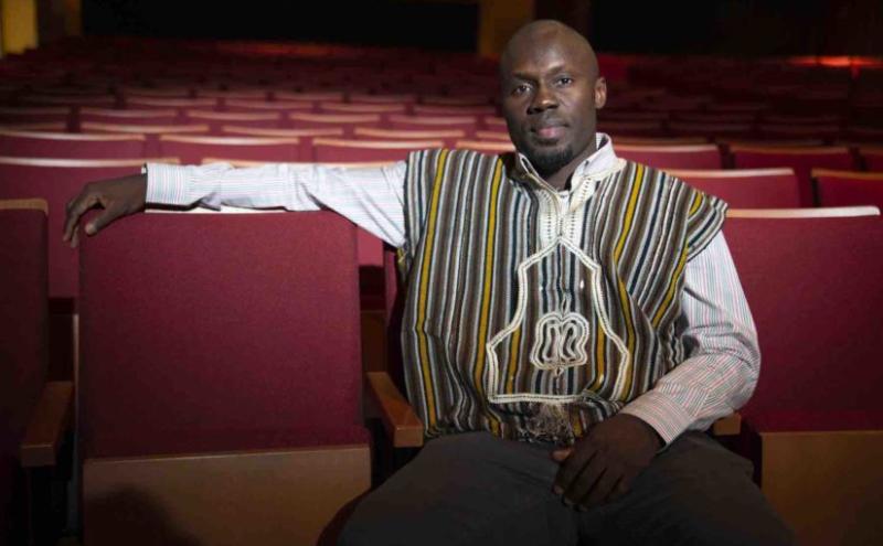 Patrick Adu sits in a red chair in a theater, wearing a striped vest.