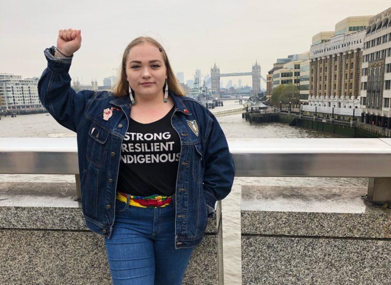 Shelby Siebers, wearing a shirt with the words, "Strong, Resilient, Indigenous," raises her fist in the air.
