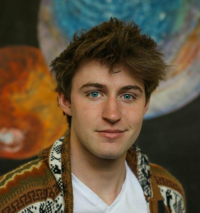 Finn Witt, wearing a brown patterned cardigan, smiles at the camera.