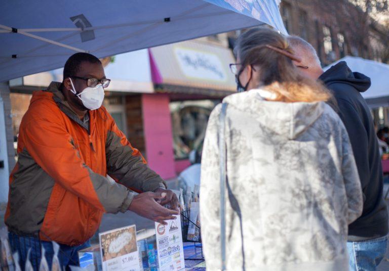 Yaw Asare, wearing an N95 mask and orange coat, interacts with a customer at his Farmers Market booth.