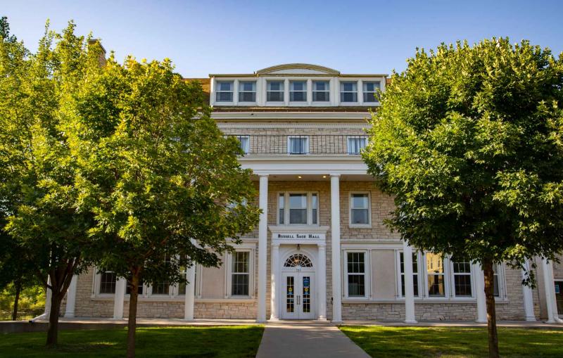 Colonial style building with stately archways and herculean pillars with brick path leading to main door flanked by trees and well-kept lawn