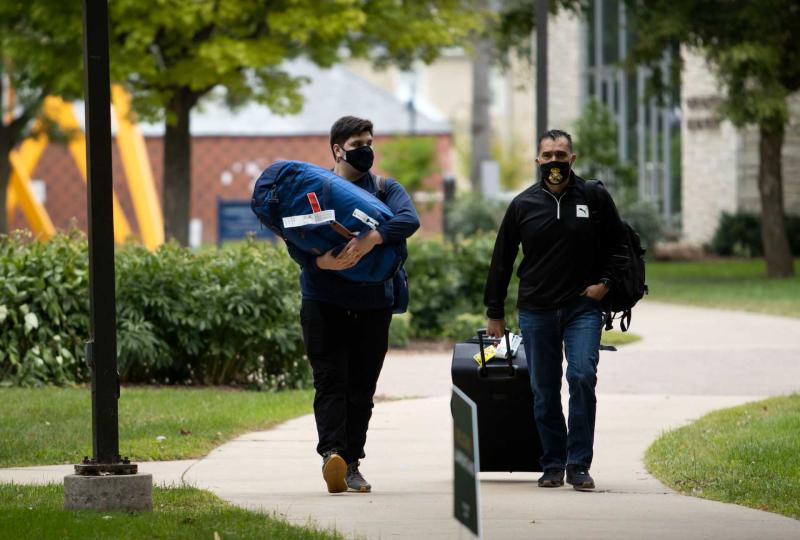 New student holds luggage, walks down campus walkway with parent pulling a suitcase