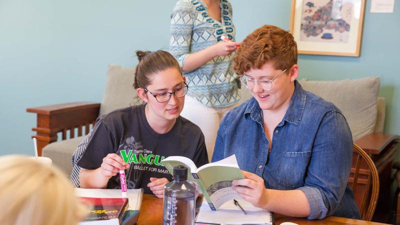 Two students seated at table looking at a shared textbook.