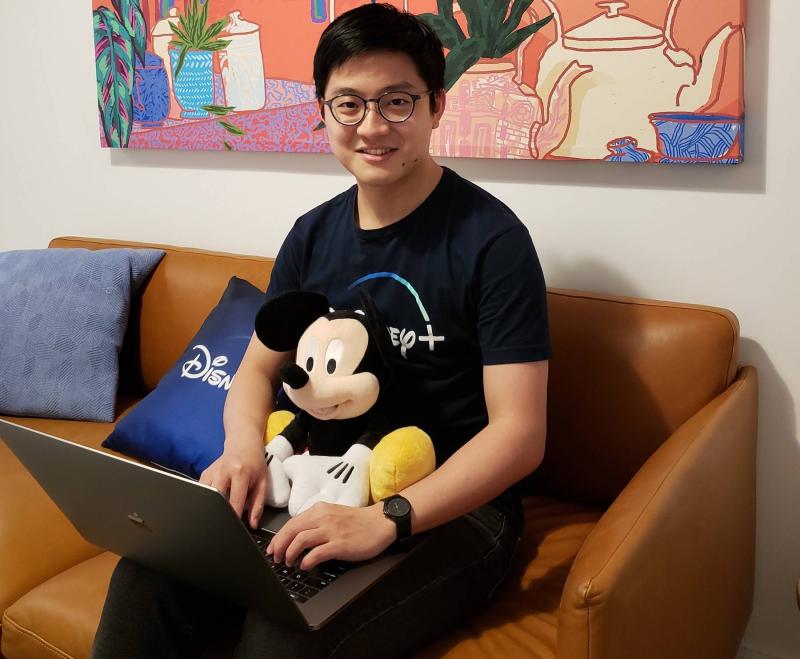 Kir Sey Fam sits on a brown leather couch with his laptop and Disney+ apparel.