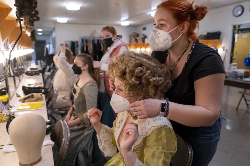 Student actors in victorian era costumes sit in front of their mirrors in the green room while other students do their hair.