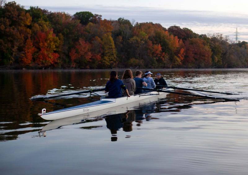 Five students row down the Fox River against fall colored leaves in background.