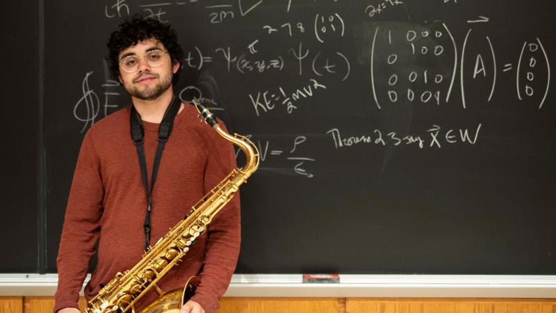 Sebastian Roman holds a saxophone in front a a blackboard depicting various mathematical formulas.