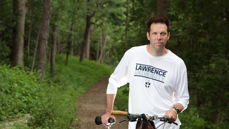 Mike O'Connor, wearing a Lawrence University shirt, stands by a bicycle on a dirt path.