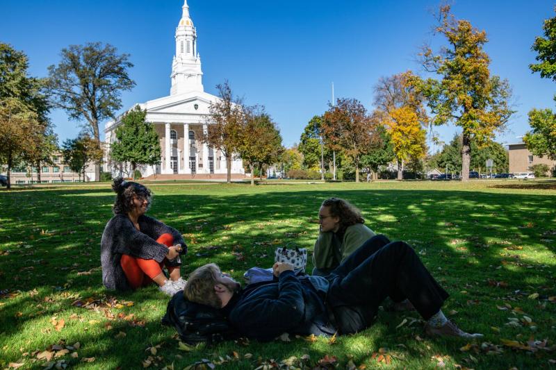 Three students sitting on Main Hall Green on sunny day with Memorial Chapel in background.