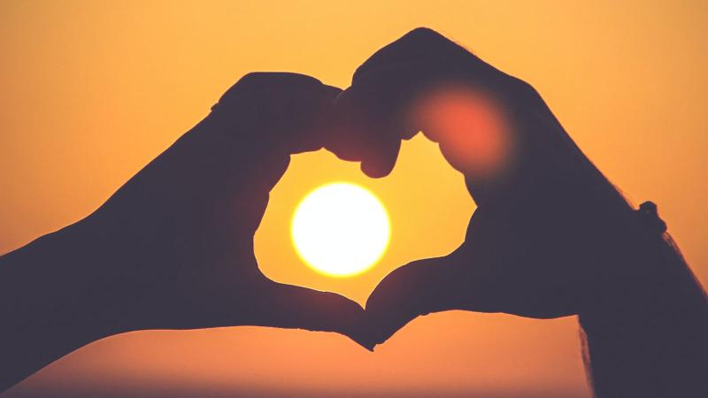 Hands making a heart shape in foreground. Sunset in background.