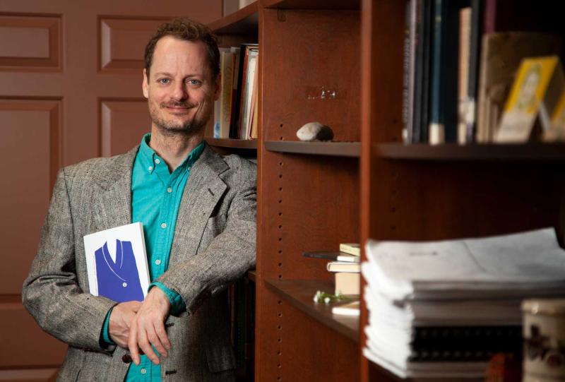 Austin Segrest holds his newly published book while leaning against a book shelf in his office.