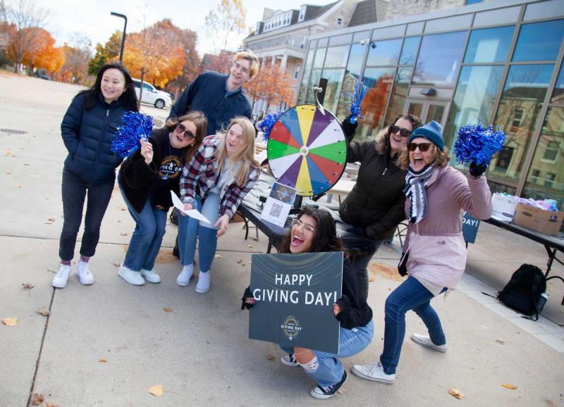Students and staff hold a Happy Giving Day sign in front of the Spin the Wheel game in front of Warch Campus Center.