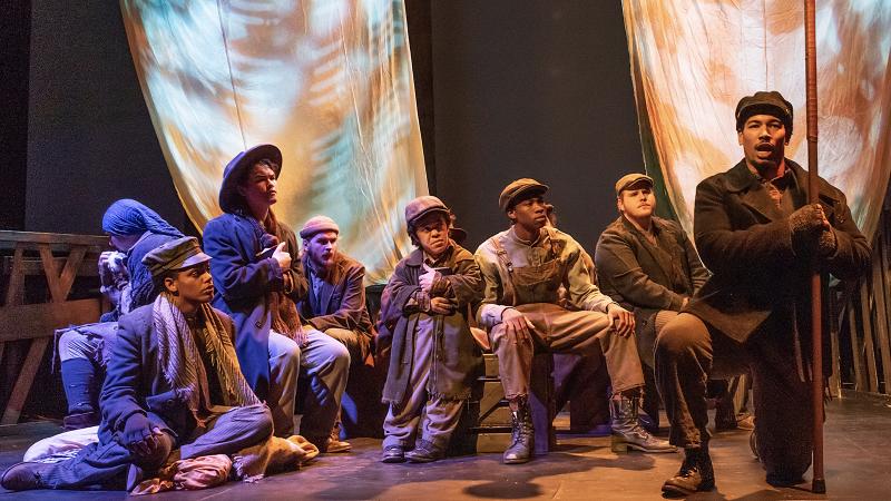 Eight members of opera cast sing while seated or kneeling onstage in atmospheric lighting