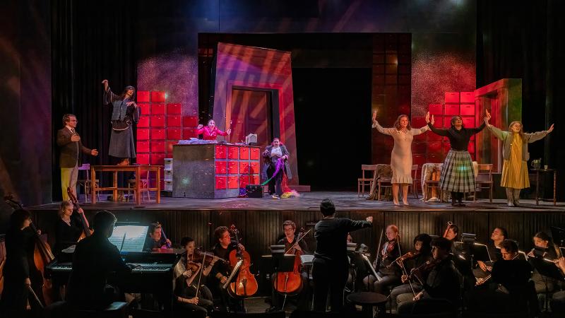 A cast of opera performers in various poses on stage while a conductor leads an orchestra in the pit below.