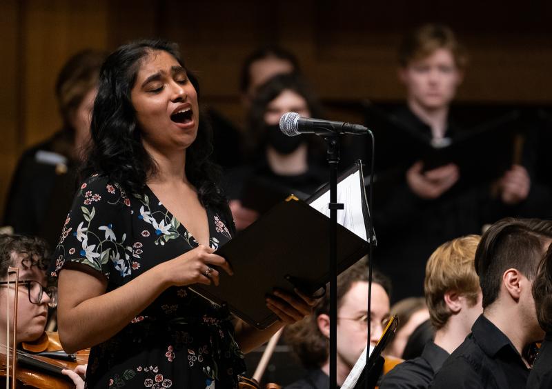 A vocalist singing a solo in performance, with an orchestra and chorus in the background.