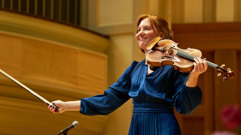 A musician holding a violin and smiling at the audience.