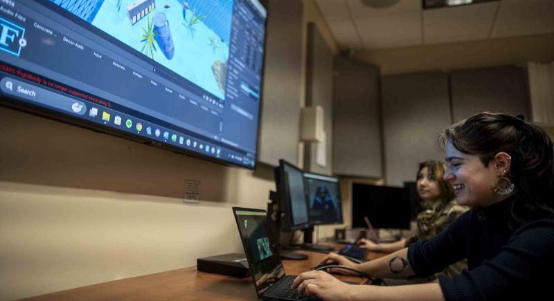 Student creating a video game world on a laptop while others watch on a big screen