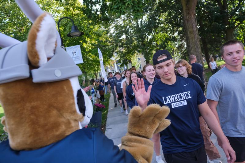 A person in a Viking mascot costume high-fives a student as a group of people walk along a tree-lined sidewalk.
