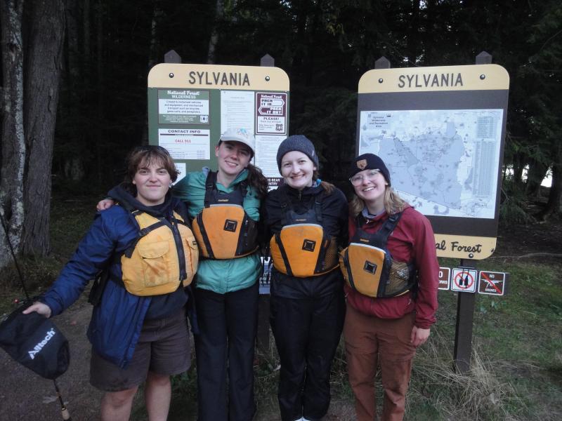 Four people wearing life jackets and outdoor gear, smiling at the camera in front of two Sylvania National Forest trailhead signs and a map, with tall trees in the background.