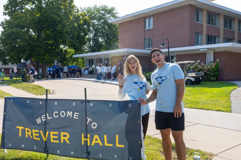 Two students stand beside a "Welcome to Trever Hall" sign, waving at the camera, while a group of people gathers near a brick residence hall in the background.
