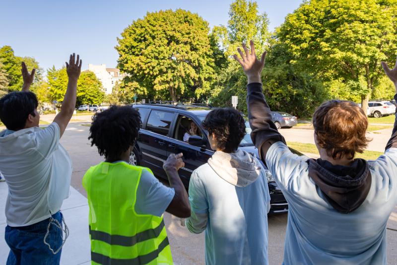Four people stand on a sidewalk with their backs to the camera, waving and cheering toward a dark minivan as it drives by on a sunny, tree-lined campus road.