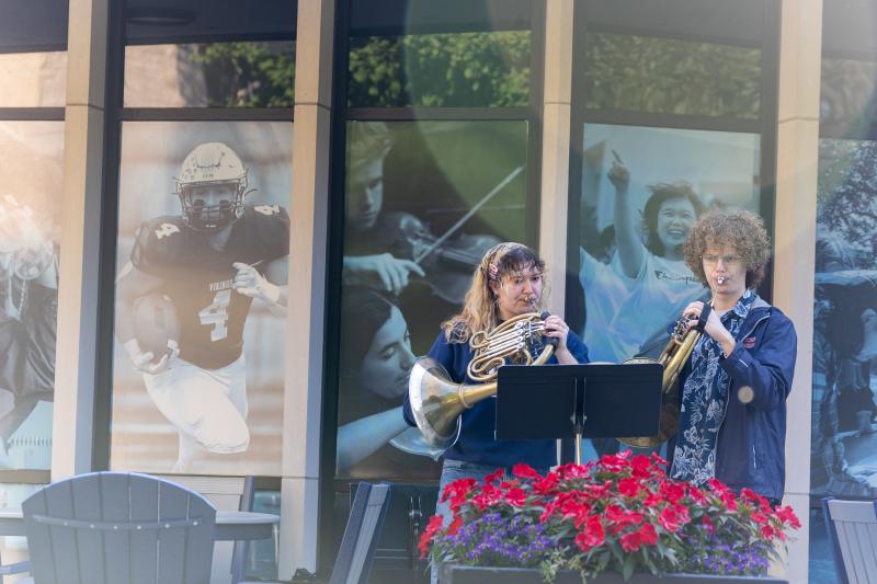 Two people play wind instruments outside in front of a building decorated with large photos of athletes, musicians, and performers. A planter with red flowers is in the foreground.