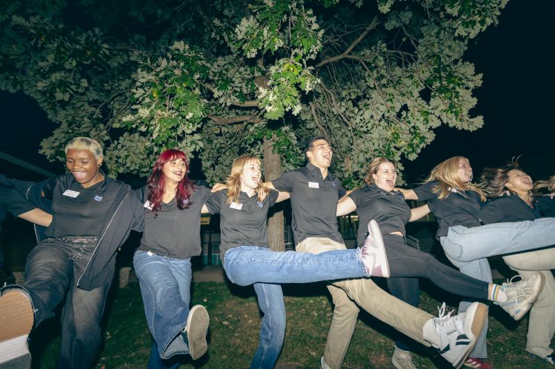 A group of six wearing matching black polo shirts standing outdoors at night under a large leafy tree, arms around each other’s shoulders, smiling as they kick one leg forward in unison.